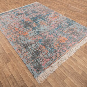 K&ouml;nnte beinhalten: Rechteckiger Teppich mit einem verwitterten, abstrakten Design. Der Teppich hat eine hellblaue Basis mit Spritzern in Grau, Orange, Rosa und T&uuml;rkis. Die Kanten haben Fransen.