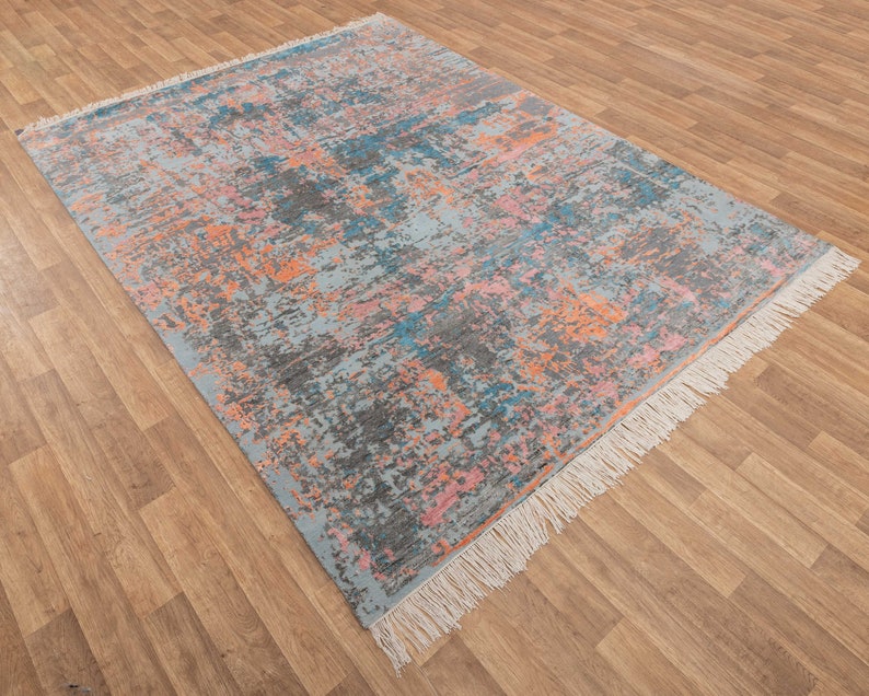K&ouml;nnte beinhalten: Rechteckiger Teppich mit einem verwitterten, abstrakten Design. Der Teppich hat eine hellblaue Basis mit Spritzern in Grau, Orange, Rosa und T&uuml;rkis. Die Kanten haben Fransen.