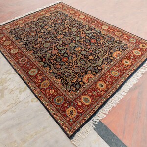May include: A traditional Persian rug with a dark blue background and intricate floral patterns in shades of red, gold, and green. The rug has a fringed border and is made of wool.