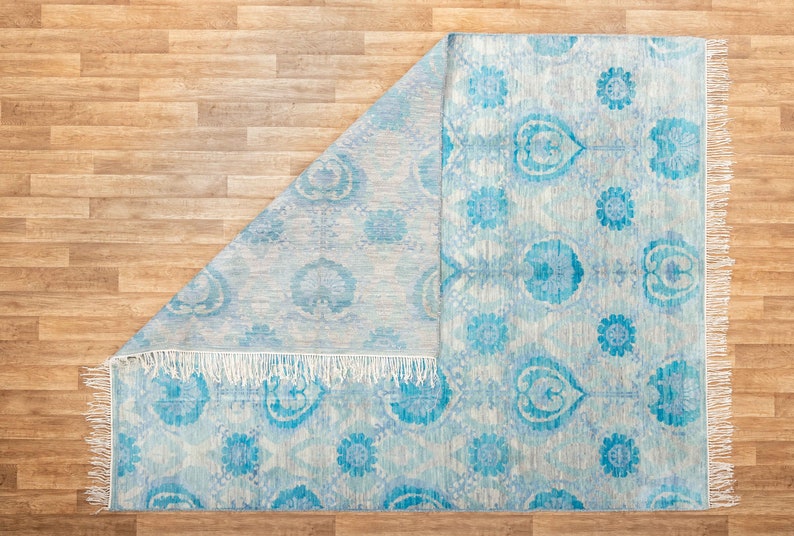 Puede incluir: Una alfombra azul claro con fondo blanco y un patr&oacute;n floral. La alfombra tiene un borde de flecos.