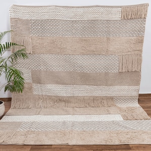 May include: A beige and white woven rug with a geometric pattern and fringe. The rug has a textured surface and is made of natural fibers.
