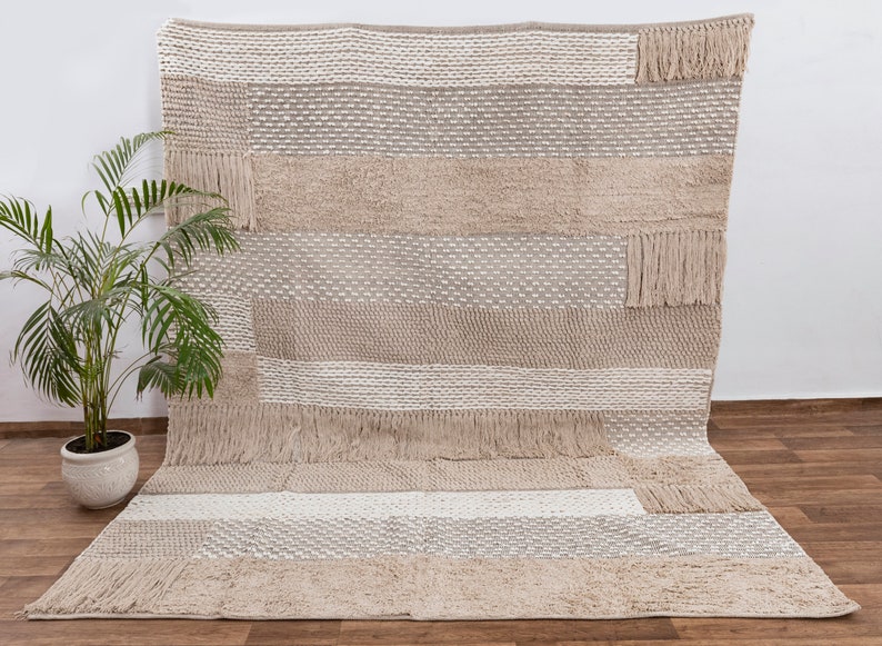 May include: A beige and white woven rug with a geometric pattern and fringe. The rug has a textured surface and is made of natural fibers.
