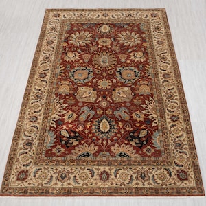 Puede incluir: Una alfombra rectangular y ornamentada con un campo central rojo intenso. La alfombra presenta un detallado patrón floral en tonos beige, azul y marrón. Un borde ancho en un beige más claro enmarca el diseño central, añadiendo a su intrincada apariencia.