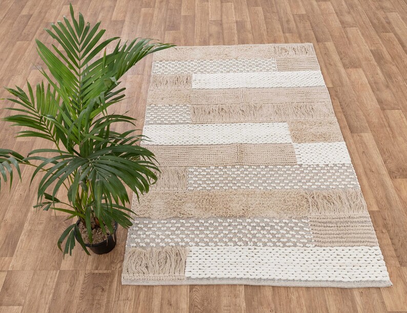 May include: A beige and white woven rug with a geometric pattern. The rug has a fringe border and is made of natural fibers.