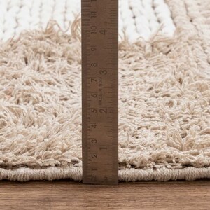 May include: Close-up of a beige and white woven rug with a fringe edge. A ruler is placed next to the rug to show the texture and detail.