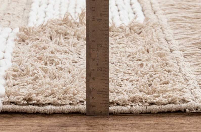 May include: Close-up of a beige and white woven rug with a fringe edge. A ruler is placed next to the rug to show the texture and detail.