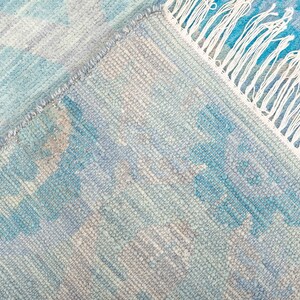 Puede incluir: Una alfombra tejida azul y blanca con un borde de flecos. La alfombra tiene un patr&oacute;n sutil de azul claro y oscuro.
