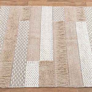 May include: A rectangular area rug with a woven design in shades of beige and white. The rug has a fringe border and a patchwork pattern.