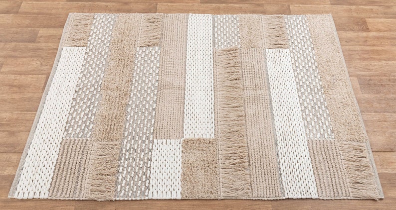 May include: A rectangular area rug with a woven design in shades of beige and white. The rug has a fringe border and a patchwork pattern.