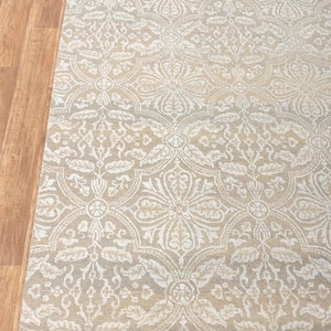 Puede incluir: Una alfombra beige con un patr&oacute;n damasco blanco. La alfombra tiene un borde de flecos.
