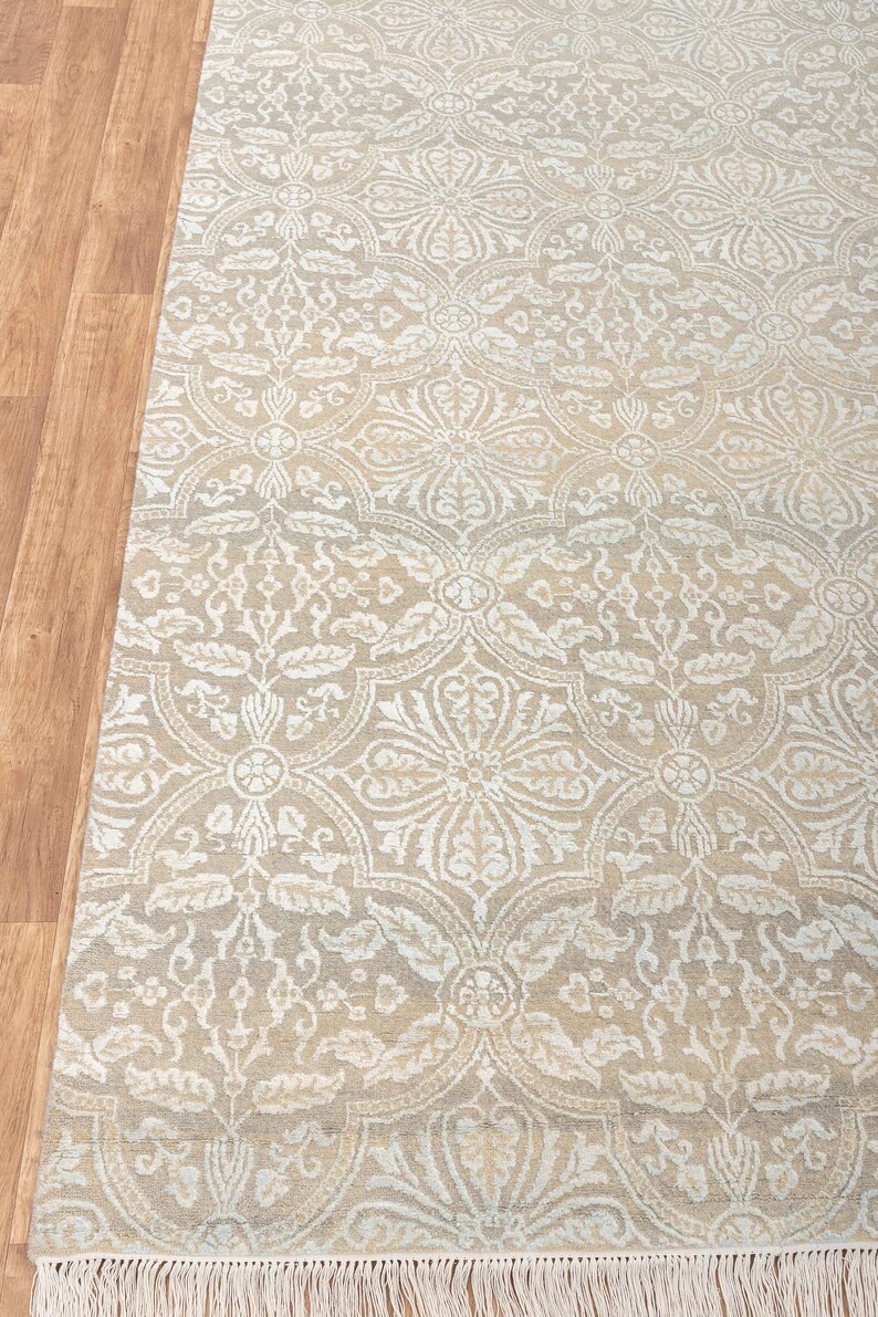 Puede incluir: Una alfombra beige con un patr&oacute;n damasco blanco. La alfombra tiene un borde de flecos.