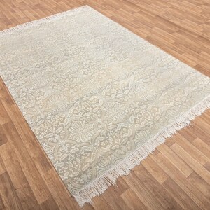 Puede incluir: Una alfombra blanca con un patr&oacute;n floral gris claro y bordes con flecos. La alfombra est&aacute; colocada sobre un suelo de madera.