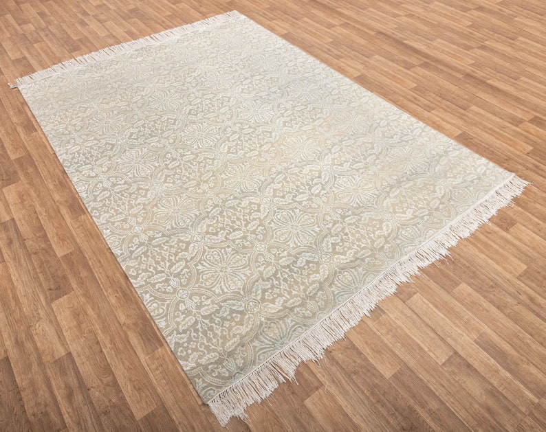 Puede incluir: Una alfombra blanca con un patr&oacute;n floral gris claro y bordes con flecos. La alfombra est&aacute; colocada sobre un suelo de madera.