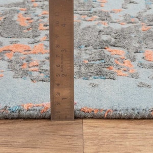 K&ouml;nnte beinhalten: Nahaufnahme eines Teppichs mit hellblauem Hintergrund und einem Muster aus grauen und korallenfarbenen Formen. Ein Lineal zeigt die Dicke des Teppichs. Das Design des Teppichs ist abstrakt und modern, geeignet f&uuml;r die Inneneinrichtung.