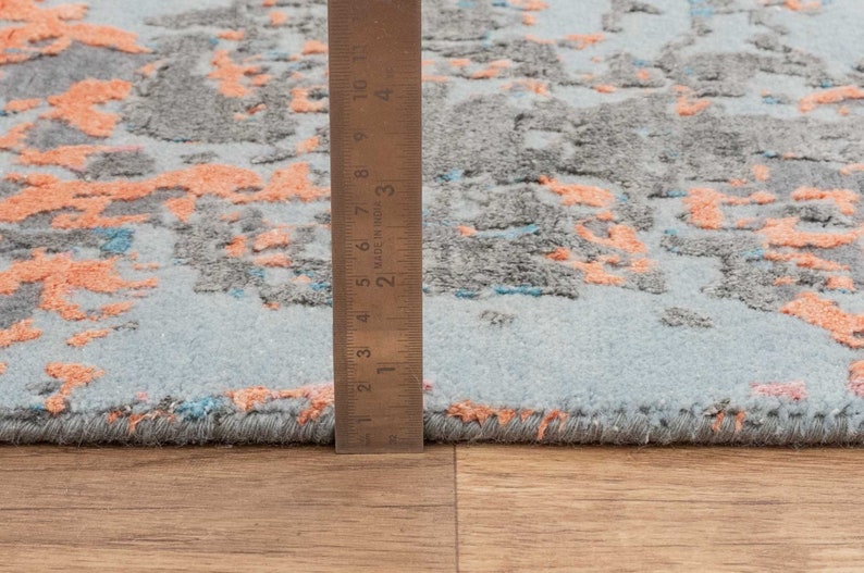 K&ouml;nnte beinhalten: Nahaufnahme eines Teppichs mit hellblauem Hintergrund und einem Muster aus grauen und korallenfarbenen Formen. Ein Lineal zeigt die Dicke des Teppichs. Das Design des Teppichs ist abstrakt und modern, geeignet f&uuml;r die Inneneinrichtung.