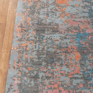 K&ouml;nnte beinhalten: Rechteckiger Teppich mit abstraktem Design. Der Teppich hat einen hellblauen Hintergrund mit grauen, orangefarbenen, rosa und blauen Flecken. Der Teppich hat einen Fransenrand und liegt auf einem Holzboden.