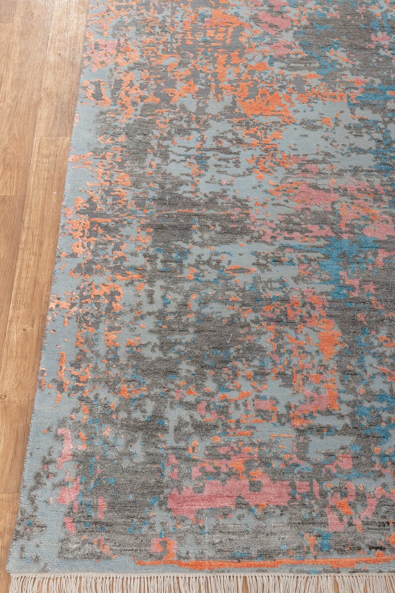 K&ouml;nnte beinhalten: Rechteckiger Teppich mit abstraktem Design. Der Teppich hat einen hellblauen Hintergrund mit grauen, orangefarbenen, rosa und blauen Flecken. Der Teppich hat einen Fransenrand und liegt auf einem Holzboden.