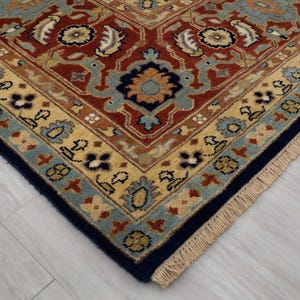 May include: Close-up of the corner of a rug with a detailed, intricate pattern. The rug has a navy blue border with a fringe and a cream, blue, and red center with floral designs.