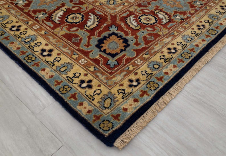 May include: Close-up of the corner of a rug with a detailed, intricate pattern. The rug has a navy blue border with a fringe and a cream, blue, and red center with floral designs.