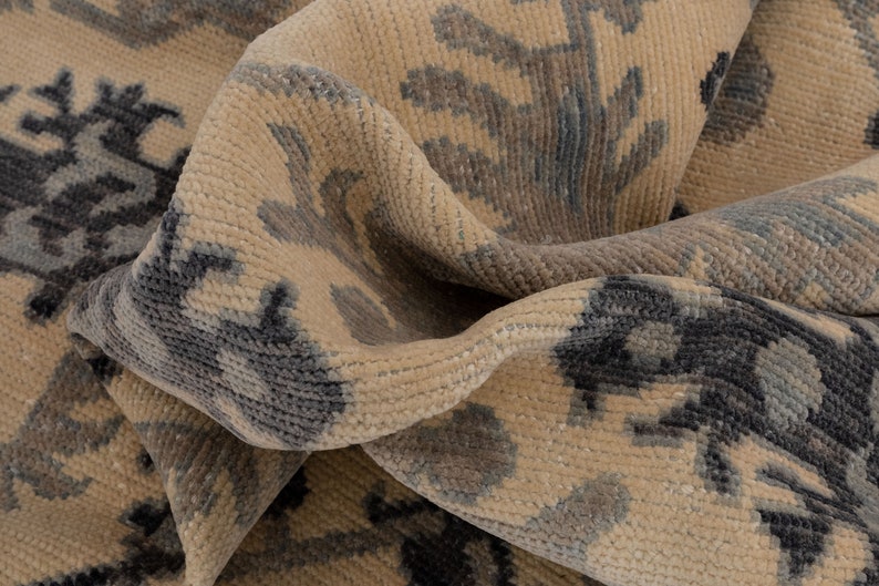 Puede incluir: Primer plano de una alfombra beige con un patr&oacute;n tejido de dise&ntilde;os florales de color gris oscuro. La alfombra est&aacute; doblada por la mitad, mostrando la textura de la tela.