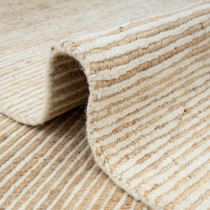 May include: Close-up of a beige and tan striped rug. The rug is made of a natural fibre material, with a rolled-up edge revealing the texture and construction. The stripes run horizontally across the rug.