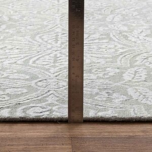 May include: A close-up of a white and gray patterned rug with a ruler measuring 6 inches. The rug has a subtle floral design.
