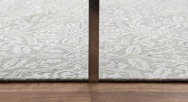 May include: A close-up of a white and gray patterned rug with a ruler measuring 6 inches. The rug has a subtle floral design.