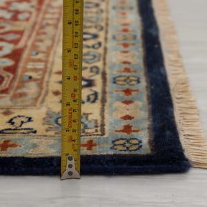 May include: Close-up of a cream-colored rug with a blue border and fringe. A measuring tape is placed along the edge of the rug, showing a measurement of 15 inches.