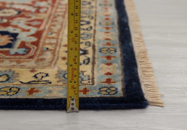May include: Close-up of a cream-colored rug with a blue border and fringe. A measuring tape is placed along the edge of the rug, showing a measurement of 15 inches.
