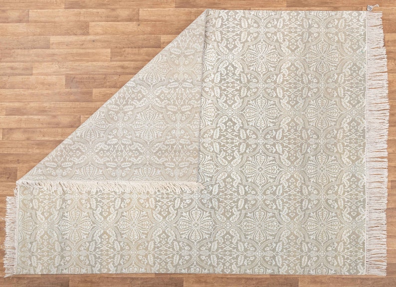 Puede incluir: Una alfombra blanca con un delicado patr&oacute;n floral y flecos en los bordes. La alfombra est&aacute; doblada por la mitad, revelando el patr&oacute;n en ambos lados.