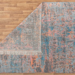 K&ouml;nnte beinhalten: Rechteckiger Teppich mit abstraktem Design. Der Teppich hat eine graue Grundfarbe mit Farbspritzern in Blau, Rosa und Orange. Die Kanten sind mit Fransen versehen. Der Teppich ist teilweise umgeklappt und zeigt die Unterseite.
