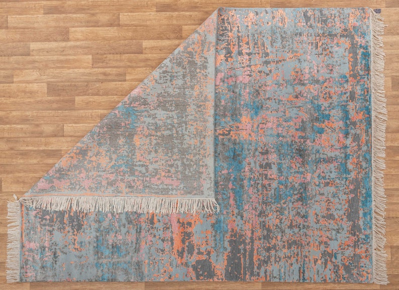 K&ouml;nnte beinhalten: Rechteckiger Teppich mit abstraktem Design. Der Teppich hat eine graue Grundfarbe mit Farbspritzern in Blau, Rosa und Orange. Die Kanten sind mit Fransen versehen. Der Teppich ist teilweise umgeklappt und zeigt die Unterseite.