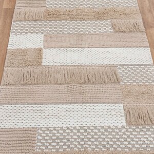 May include: A beige and white woven rug with a geometric pattern and fringe trim. The rug has a textured surface and is made of natural fibers.