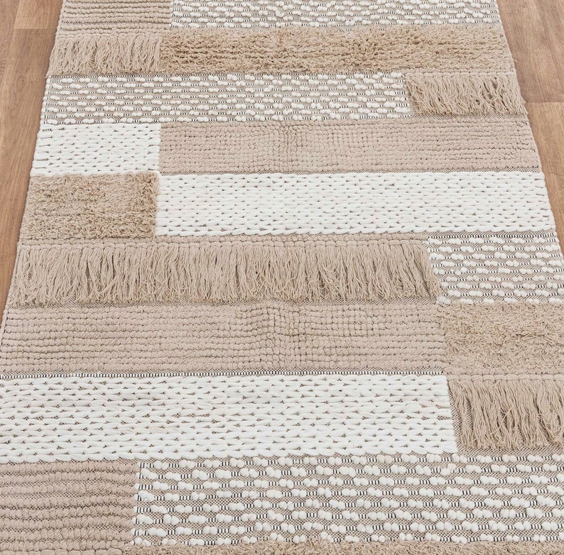May include: A beige and white woven rug with a geometric pattern and fringe trim. The rug has a textured surface and is made of natural fibers.