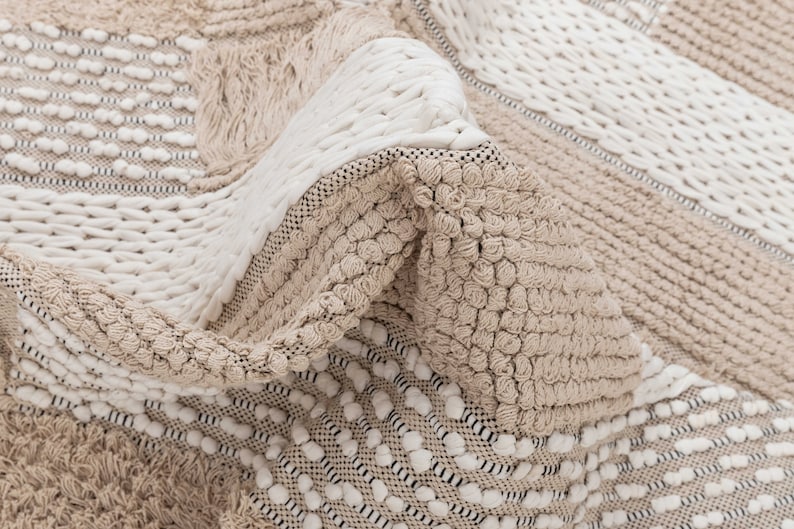 May include: A close-up of a beige and white woven rug with a textured, bumpy surface. The rug has a geometric pattern with different sized squares and rectangles.