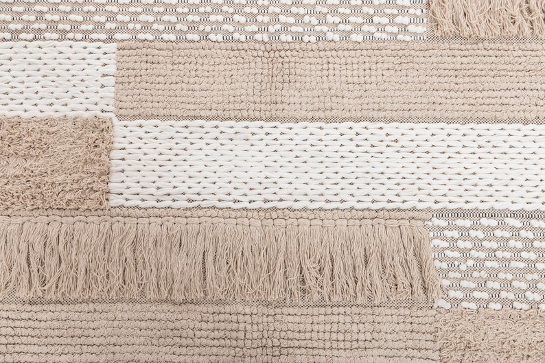 May include: A beige and white woven rug with a patchwork design. The rug has a fringe border and is made of different textures.