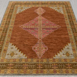 Puede incluir: Una alfombra de color óxido con un patrón de diamante en tonos de rosa, beige y marrón. La alfombra tiene un borde con intrincados diseños florales y geométricos en tonos de oro, marrón y beige.