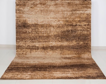 May include: A large, rectangular area rug with a textured, abstract design. The rug features a blend of brown and beige tones, creating a layered effect. The rug is rolled out on a white floor, with the top portion against a white wall.