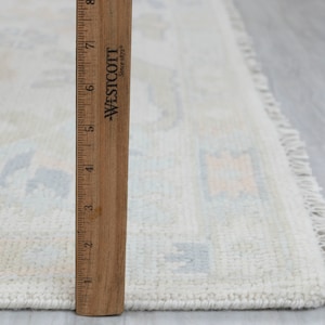 May include: A white rug with a light blue and peach geometric pattern. The rug has a fringe border and is measured against a wooden ruler.