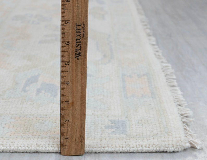 May include: A white rug with a light blue and peach geometric pattern. The rug has a fringe border and is measured against a wooden ruler.