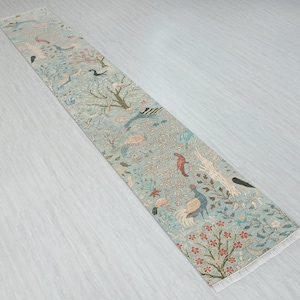 Könnte beinhalten: Ein langer, schmaler Teppich mit hellblauem Hintergrund und einem detaillierten Muster aus Vögeln, Bäumen und floralen Elementen in Rosa-, Grün- und Brauntönen. Der Teppich hat einen Fransenrand.