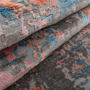 K&ouml;nnte beinhalten: Nahaufnahme eines aufgerollten Teppichs mit abstraktem Design. Der Teppich hat einen grauen Hintergrund mit Spritzern in Rosa, Orange und Blau. Die Textur wirkt weich und pl&uuml;schig, was auf einen komfortablen Bodenbelag hindeutet.
