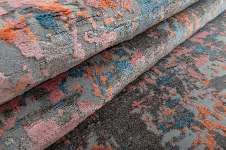 K&ouml;nnte beinhalten: Nahaufnahme eines aufgerollten Teppichs mit abstraktem Design. Der Teppich hat einen grauen Hintergrund mit Spritzern in Rosa, Orange und Blau. Die Textur wirkt weich und pl&uuml;schig, was auf einen komfortablen Bodenbelag hindeutet.