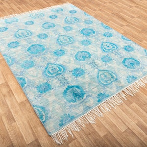 Puede incluir: Una alfombra azul claro con fondo blanco y un patr&oacute;n floral. La alfombra tiene un borde de flecos.