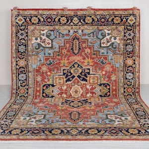 Puede incluir: Una alfombra rectangular grande con un diseño simétrico intrincado. La alfombra presenta un medallón central en tonos de rojo, azul y dorado, rodeado por un borde con patrones florales y geométricos. La alfombra se exhibe sobre un suelo claro.