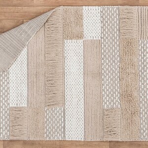 May include: A beige and white woven rug with a geometric pattern. The rug has a fringe border and is made of natural fibers.