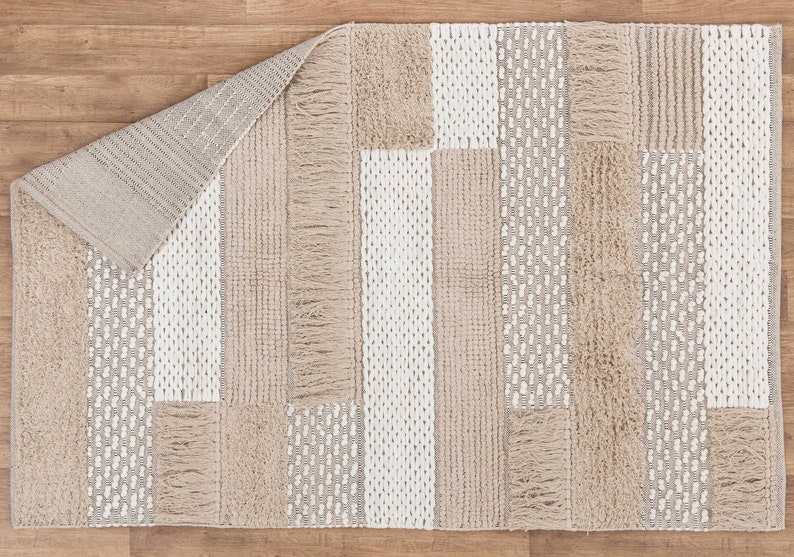 May include: A beige and white woven rug with a geometric pattern. The rug has a fringe border and is made of natural fibers.