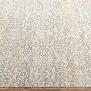 Puede incluir: Una alfombra beige con un intrincado patr&oacute;n floral blanco. La alfombra tiene un brillo sutil y una textura suave.