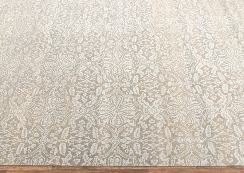 Puede incluir: Una alfombra beige con un intrincado patr&oacute;n floral blanco. La alfombra tiene un brillo sutil y una textura suave.
