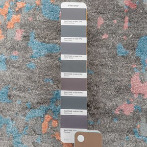 K&ouml;nnte beinhalten: Ein grauer Teppich mit abstrakten Mustern in Blau, Orange und Rosa. Rechts befindet sich eine Pantone-Farbkarte mit verschiedenen Graut&ouml;nen. Das Design des Teppichs weist ein strukturiertes, gesprenkeltes Aussehen auf.